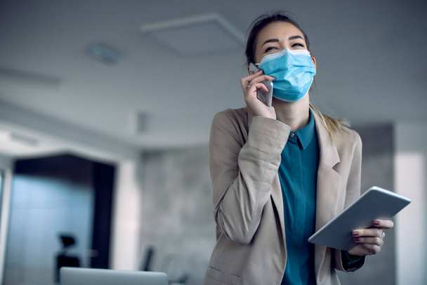 Young businesswoman having fun while communicating over cell phone and working on digital tablet in the office during coronavirus pandemic.  - 写真・画像