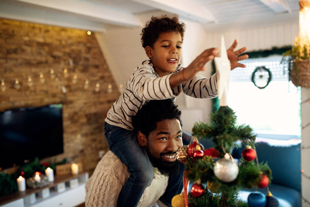 Ευτυχισμένος Αφρο-Αμερικανός πατέρας κουβαλάει το γιο του στους ώμους και τον βοηθάει να βάλει αστέρι πάνω στο χριστουγεννιάτικο δέντρο..  - Φωτογραφία, εικόνα