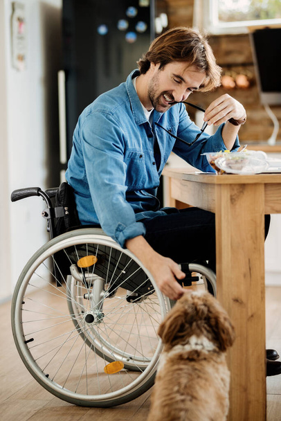 Happy man in wheelchair enjoying with his dog while having a meal at home.  - Photo, Image