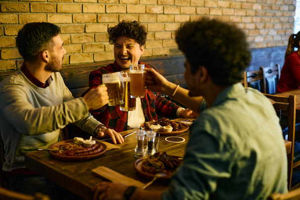 Νεαρός άντρας γελάει και κάνει πρόποση με μπύρα με τους άντρες φίλους του ενώ τρώει σε μια παμπ.. - Φωτογραφία, εικόνα