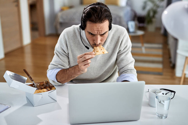 Молодий підприємець використовує ноутбук і їсть під час роботи вдома
.  - Фото, зображення