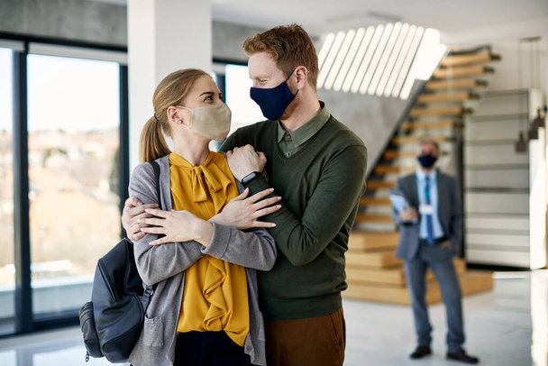 Young embraced couple wearing protective face masks and looking at each other while buying new apartment. Real estate agent is in the background.  - Foto, afbeelding