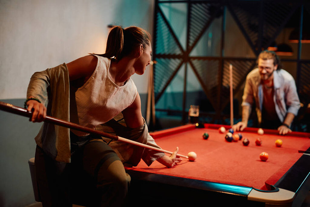 Young woman aiming at white ball while playing snooker with her boyfriend in pool hall. - Foto, Imagen