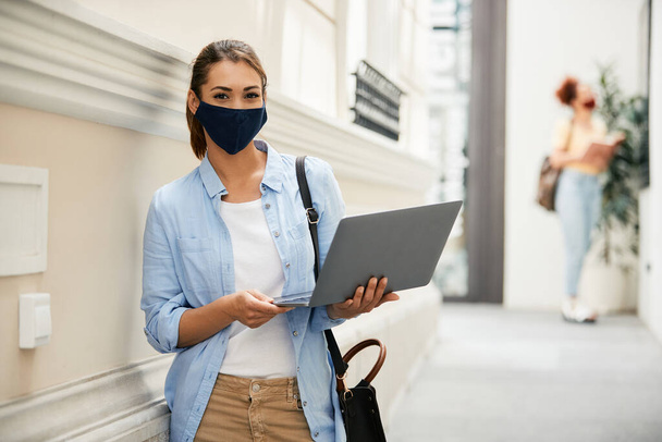 Happy female college student using laptop in a hallway while waring protective face mask due to COVID-19 pandemic and looking at camera. - Photo, image