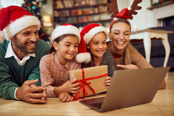 Happy family showing wrapped Christmas present while using laptop and talking to someone via video call at home. - Fotografie, Obrázek