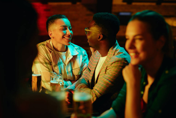 Happy Black woman communicating with her boyfriend while drinking beer in a pub at night.  - 写真・画像