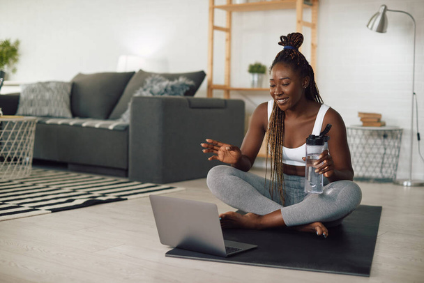 Happy Afro-Amerikaanse vrouw met water pauze tijdens online oefening klasse over laptop thuis. - Foto, afbeelding