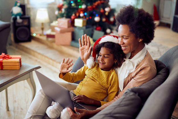 Happy black mother and daughter using laptop and waving during video call on Christmas at home. - Fotó, kép