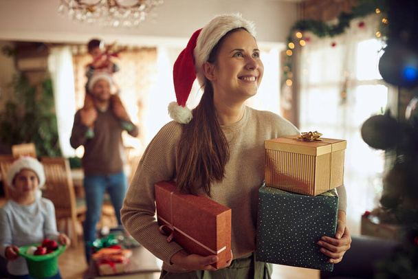 Счастливая женщина держит завернутые подарки во время празднования Рождества со своей семьей дома. - Фото, изображение