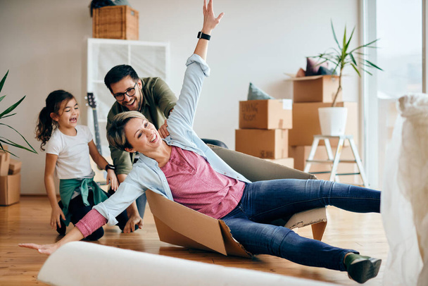 Happy woman with arms outstretched having fun while being pushed by her husband and daughter in cardboard box at their new home.  - Zdjęcie, obraz
