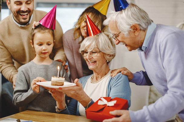Счастливая расширенная семья празднует день рождения пожилой женщины и удивляет ее тортом и подарками. Сосредоточься на пожилой женщине.  - Фото, изображение