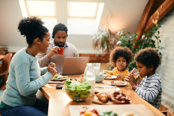 Boldog afro-amerikai anya beszél a gyerekeivel reggeli közben az asztalnál, míg apa a laptopon dolgozik.. - Fotó, kép
