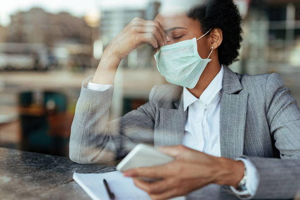 Exhausted African American businesswoman wearing protective face mask and holding her head in pain. The view is through the glass.  - Photo, image