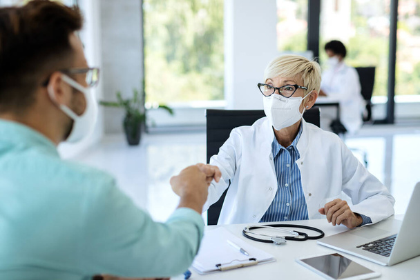 Mature doctor and her patient wearing protective face masks while greeting with fists at medical clinic.  - 写真・画像
