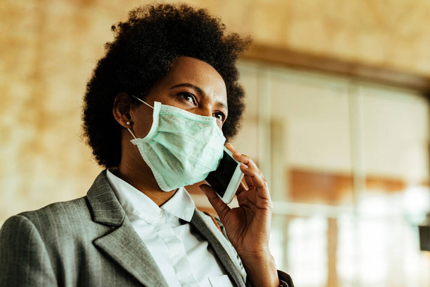 Low angle view of black businesswoman wearing protective mask during virus epidemic and communicating on mobile phone while being at airport corridor.  - Foto, afbeelding