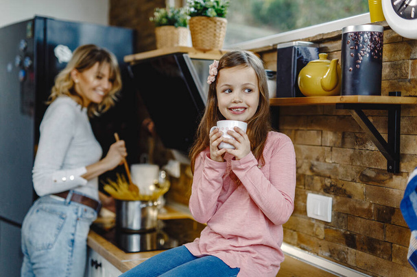 Smiling girl enjoying in cup of tea while her mother in preparing food in the kitchen.  - Fotoğraf, Görsel
