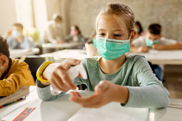 Elementary student wearing protective face mask and cleaning her hands with a disinfectant in the classroom.  - Foto, afbeelding