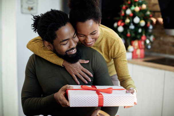 Glückliche schwarze Frau umarmt ihren Mann und schenkt ihm zu Weihnachten zu Hause ein Geschenk.  - Foto, Bild
