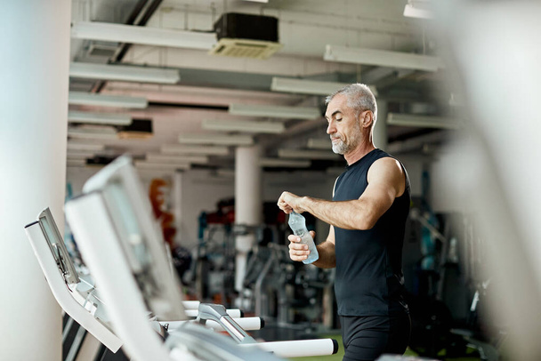 Mature athletic man drinks water after running on treadmill in g - Photo, Image