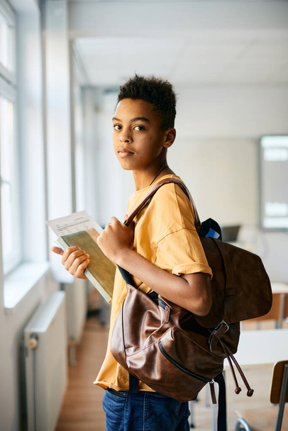 Афроамериканский школьник с рюкзаком и книгами, стоящими в классе и смотрящими в камеру.  - Фото, изображение