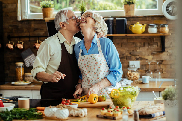 Щасливе подружжя старших осіб, яке разом готує їжу. Чоловік цілує свою дружину..  - Фото, зображення