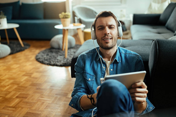Junger Mann nutzt digitales Tablet und genießt Musik über Kopfhörer, während er sich im Wohnzimmer entspannt.  - Foto, Bild