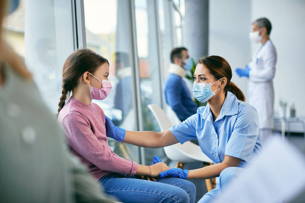 Gelukkig verpleegster houdt hand in hand met een klein meisje terwijl ze met haar communiceert in een lobby in de medische kliniek. Ze dragen beschermende gezichtsmaskers als gevolg van COVID-19 pandemie.  - Foto, afbeelding