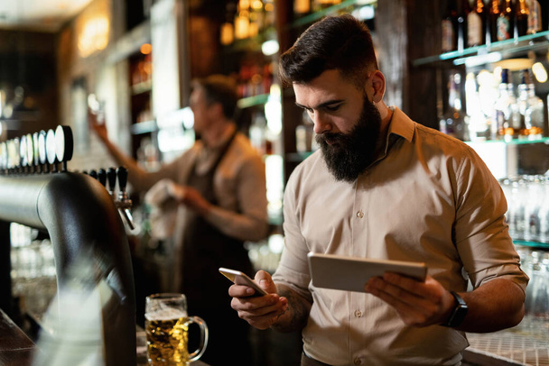 Young barista texting on smart phone while using touchpad in a bar.  - Foto, Imagen