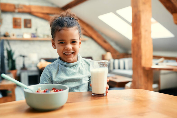 Happy African American little girl having milk mustache while eating breakfast at dining table and looking at camera. - Foto, Imagem