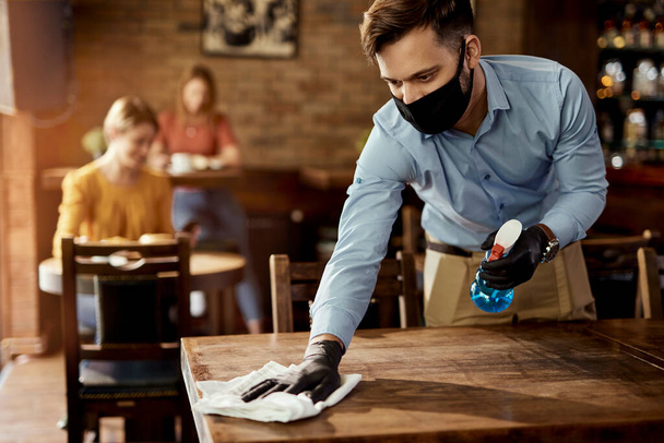 Молодой официант в защитной маске во время уборки столов во время работы в кафе.  - Фото, изображение
