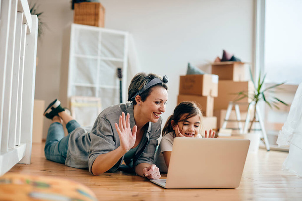 Happy single mother and daughter using laptop and greeting someone during video call while relaxing on the floor at their new home.  - Fotografie, Obrázek