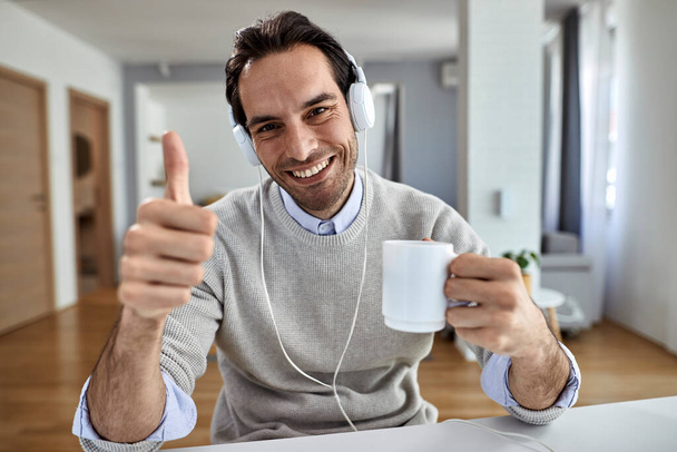 Νεαρός ευτυχισμένος επιχειρηματίας πίνοντας καφέ και δείχνοντας αντίχειρες επάνω, ενώ εργάζονται στο σπίτι.  - Φωτογραφία, εικόνα
