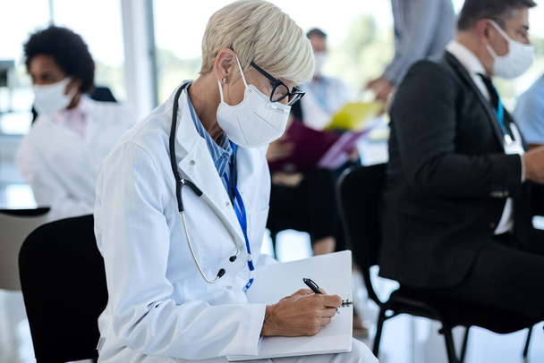 Зрілий медичний працівник носить захисну маску для обличчя і пише в записнику, відвідуючи семінар у залі засідань
.  - Фото, зображення
