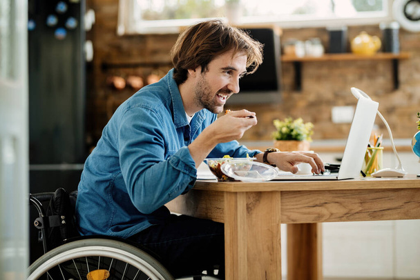 Jong gelukkig man in rolstoel eten tijdens het gebruik van laptop thuis.  - Foto, afbeelding