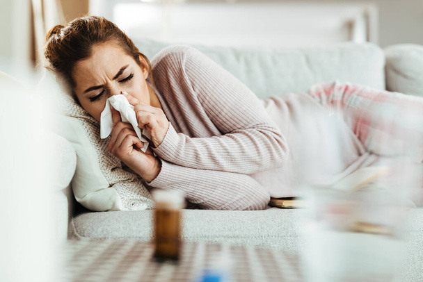 Young woman with flu virus lying down on sofa and blowing her nose.  - Photo, image