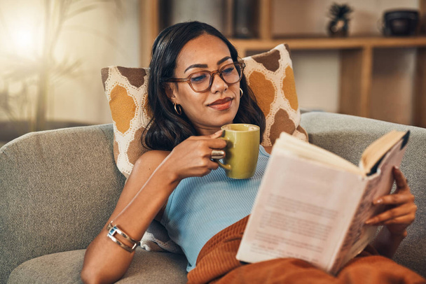 Piękna mieszana kobieta rasy czytania książki i picia kawy w salonie w domu. Hiszpanie leżą samotnie na sofie i cieszą się powieścią. Poczucie relaksu w weekend z fabułą i herbatą. - Zdjęcie, obraz