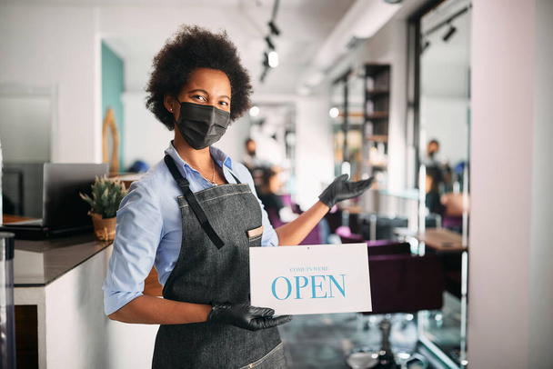 African American hairdresser wearing face mask while holding open sign and welcoming her customers at hair salon. - Foto, Bild