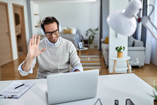 Молодой счастливый бизнесмен машет рукой во время онлайн-встречи за ноутбуком из дома. - Фото, изображение