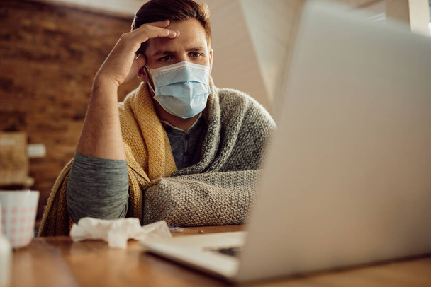 Distraught man using computer while wearing face mask and sitting wrapped in a blanket at home. - Foto, Imagem