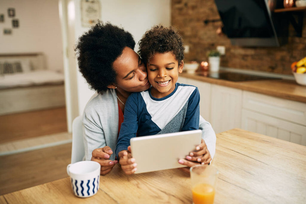 Loving African American mother kissing her son who is sitting on her lap and surfing the net on touchpad.  - Valokuva, kuva