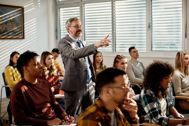 Mature professor pointing at something while holding lecture to his students in the classroom.  - Fotoğraf, Görsel