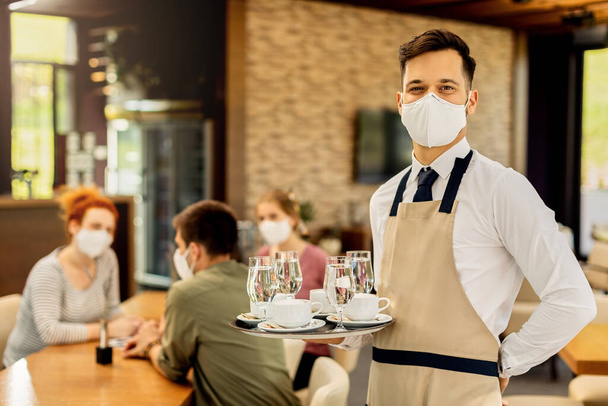 Portrait of happy waiter with protective face mask serving customers in a cafe and looking at camera.  - Fotoğraf, Görsel