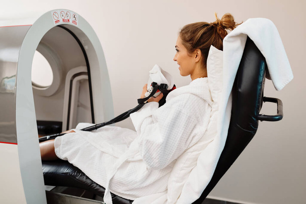 Young happy woman receiving oxygen therapy in hyperbaric chamber at health spa. - Фото, изображение