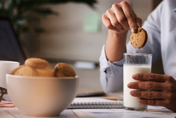 Close-up of freelance worker having a break and dipping cookie in a glass of milk.  - Fotó, kép