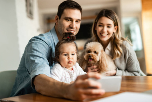 Young happy family with a dog using smart phone while taking selfie at home. Focus is on girl.  - Fotó, kép
