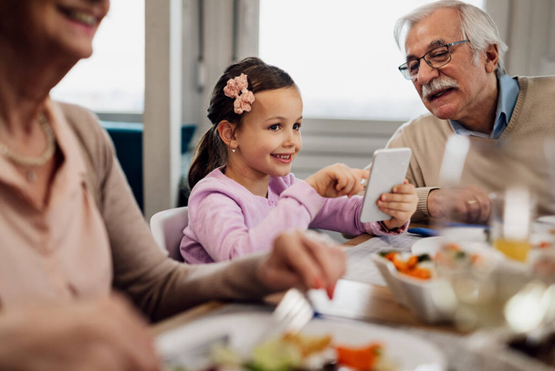 Happy little girl having lunch with her grandparents and using smart phone at dining table.  - Фото, изображение
