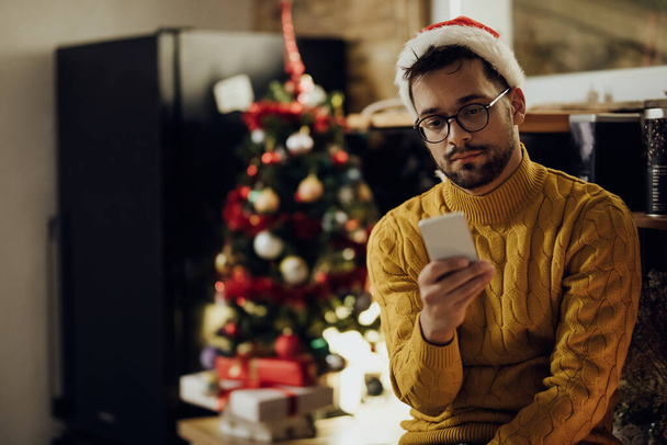 Молодой грустный мужчина пишет смс на смартфоне, чувствуя себя одиноким в канун Рождества дома. - Фото, изображение