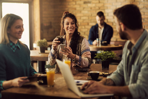 Молода щаслива жінка п'є каву і смс на мобільний телефон, розмовляючи зі своїми друзями в кафе
.  - Фото, зображення