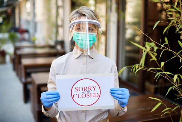 Waitress with protective face mask and visor holding closed sign at outdoor cafe.  - Foto, imagen