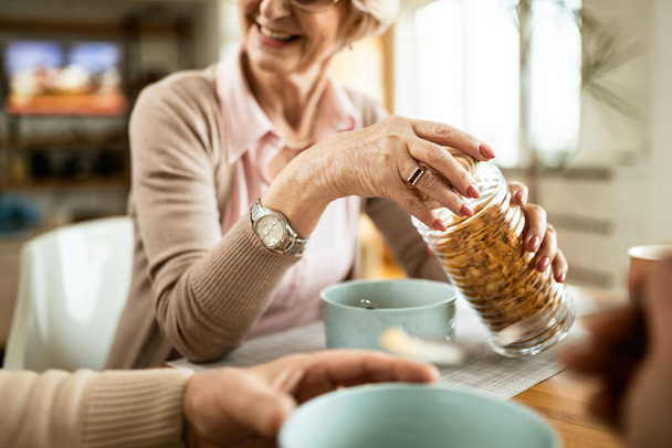 Close-up of mature woman opening jar with corn flakes while eating breakfast with her husband at home.  - Zdjęcie, obraz